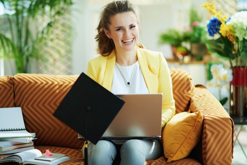 Girl wearing yellow sitting on a couch working on a laptop with a grad cap