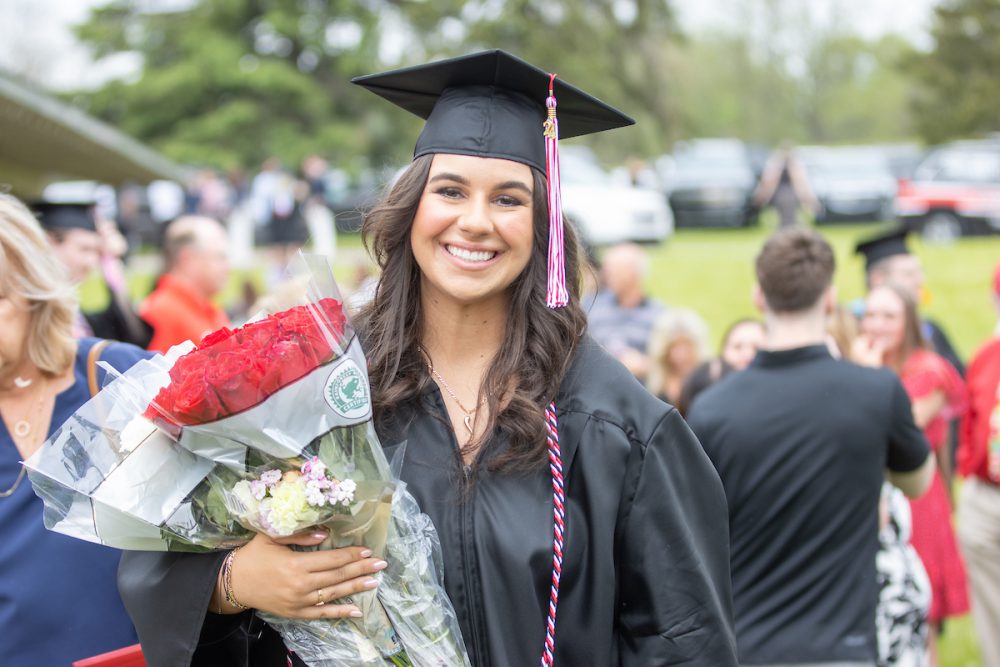 Photo of smiling female CUAA graduate in cap and gown holding a bouquet of flowers